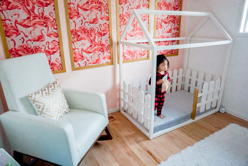 girls nursery room: flamingo wallpaper with gold frame in modern chinoiserie on a pink wall, white modern rocking chair, and white wooden house bed