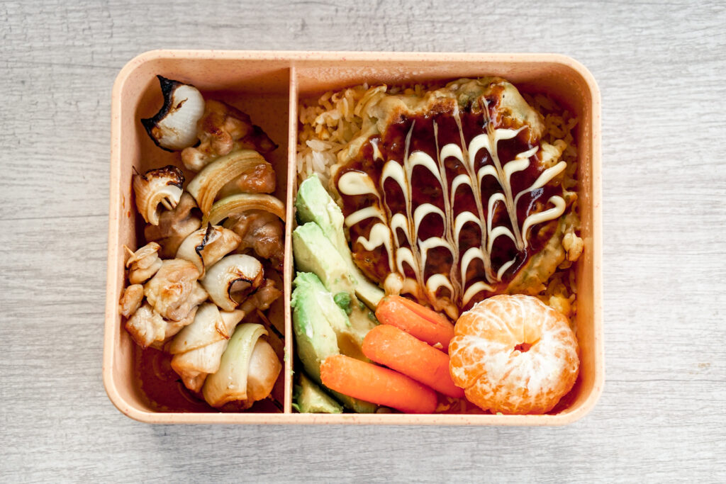 Japanese bentou with okonomiyaki, yakitori, avocado, carrots, and a satsuma orange