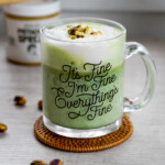 pistachio matcha latte front view in glass mug with matcha whisk and pistachio butter in background